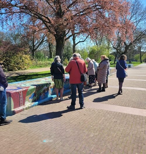 Groep mensen staat buiten op een zonnige dag, kijkend naar kleurrijke objecten. Bomen met roze bloesem op de achtergrond.