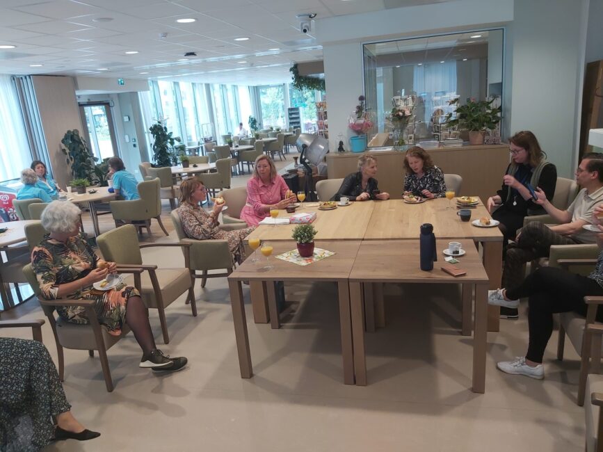 Groep medewerkers zit aan een tafel in een lichte ruimte voor een lunch, omringd door planten en grote ramen.