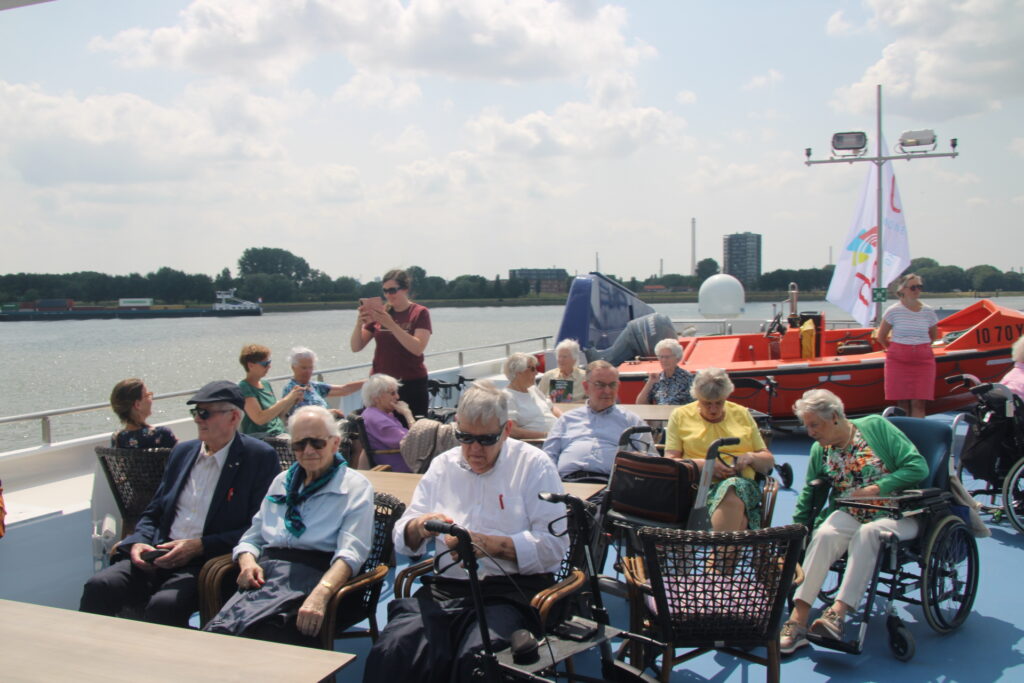 Groep ouderen geniet van een zonnige dag op een boot, zittend in rolstoelen en stoelen, met een rivier en stad op de achtergrond.