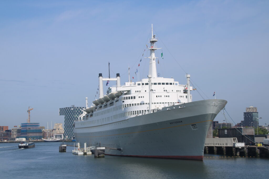 Wit passagiersschip aangemeerd in een haven met moderne gebouwen op de achtergrond en een heldere blauwe lucht erboven.