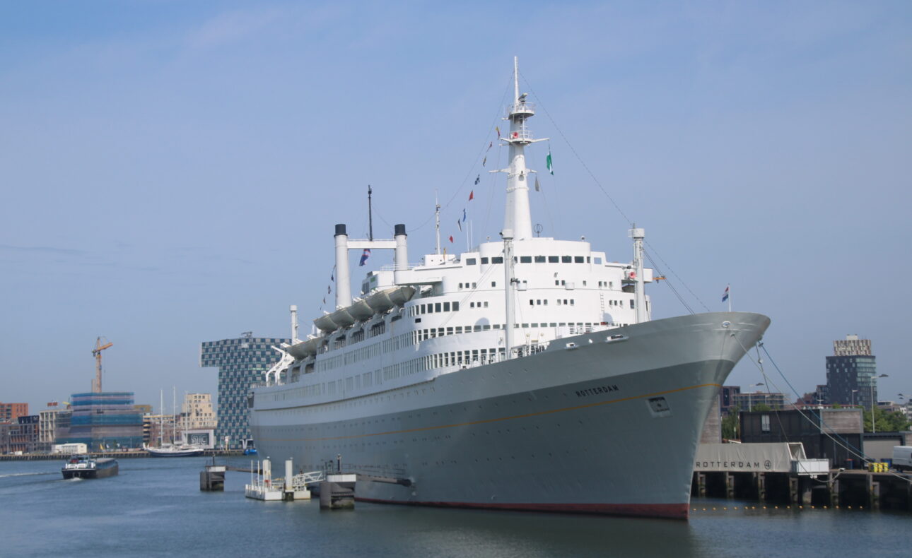 Wit passagiersschip aangemeerd in een haven met moderne gebouwen op de achtergrond en een heldere blauwe lucht erboven.