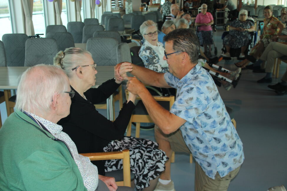 Een man in een blauw overhemd danst met een oudere vrouw in een rolstoel in een zaal vol met andere ouderen.