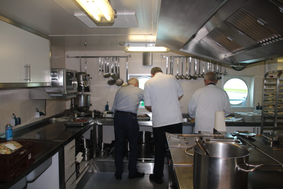 Drie koks werken samen in een professionele keuken. Ze bereiden gerechten voor op een groot fornuis onder fel licht.