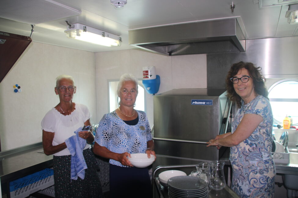 Drie vrouwen staan in een keuken en doen de afwas. Ze lachen terwijl ze borden en glazen schoonmaken bij een vaatwasser.