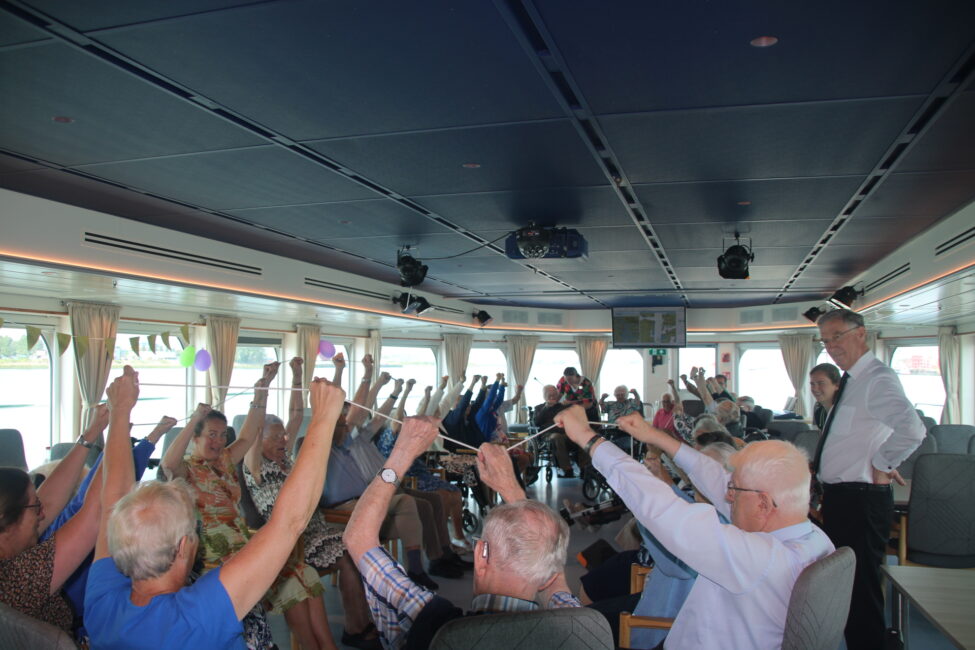 Groep ouderen in een zaal met blauwe plafonds, zittend in stoelen, steekt gezamenlijk hun armen omhoog tijdens een activiteit.