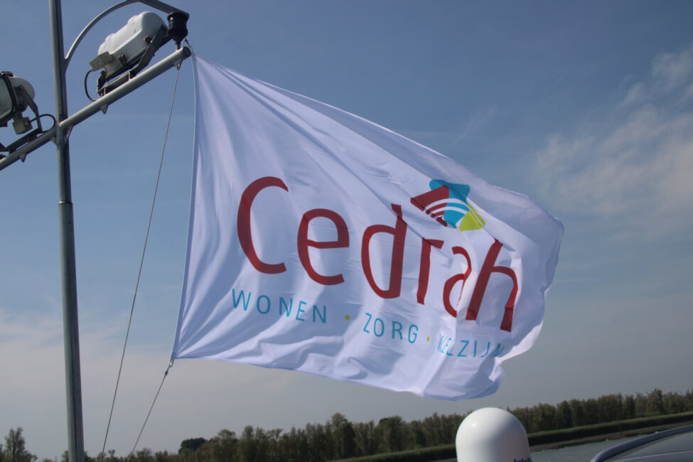 Witte vlag met het logo van Cedrah wappert in de wind tegen een blauwe lucht. Onder het logo staan de woorden "Wonen Zorg Zorg".