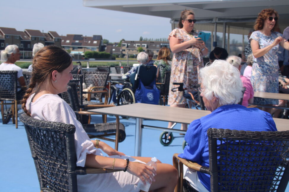 Groep mensen, waaronder ouderen, zitten buiten op een terras. Ze praten en genieten van het zonnige weer.