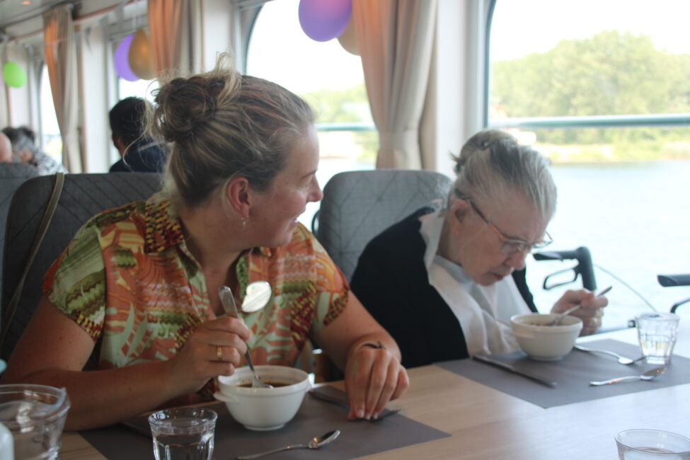Twee vrouwen zitten aan tafel op een boot en eten soep. De vrouw links kijkt naar de vrouw rechts, die in een rolstoel zit.