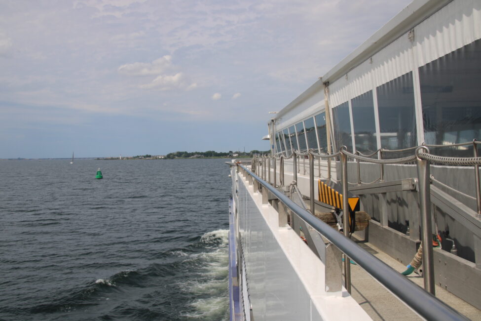 Zijaanzicht van een varende veerboot op een rustige zee, met een blauwe lucht en een kustlijn in de verte aan de horizon.