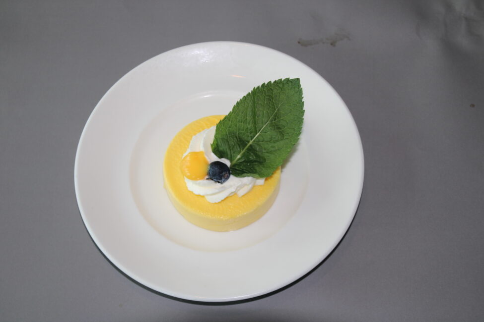 Een dessert op een wit bord, met een gele mousse, slagroom, een groene blaadje en een blauwe bes als garnering.