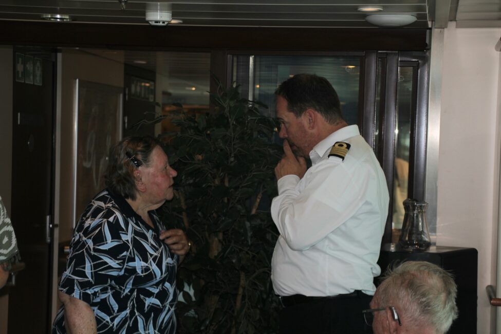 Een vrouw in gesprek met een man in uniform in een kamer met planten. Beiden lijken aandachtig te luisteren naar elkaar.