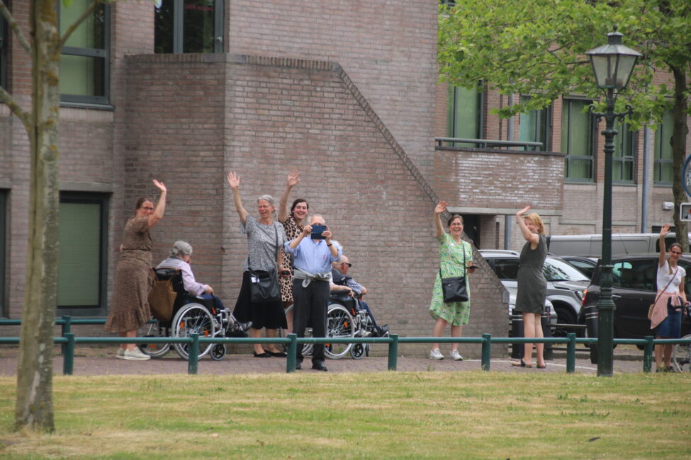 Groep mensen zwaait naar de camera buiten een gebouw. Enkele personen zitten in rolstoelen, anderen staan ernaast.