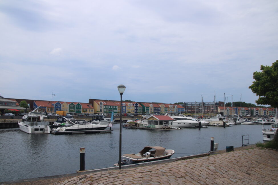 Haven met aangemeerde boten en kleurrijke huizen op de achtergrond, onder een bewolkte hemel. Een lantaarnpaal staat op de kade.