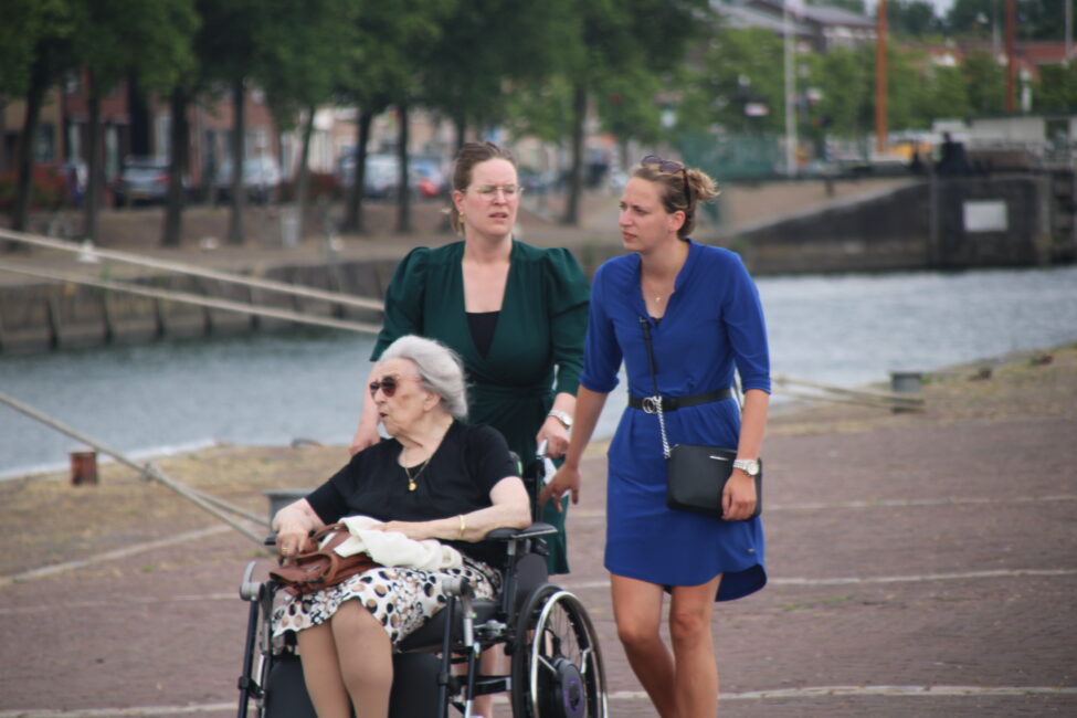 Drie vrouwen wandelen langs een rivier; een oudere vrouw zit in een rolstoel, begeleid door twee jongere vrouwen.