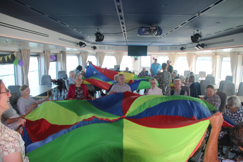 Groep ouderen speelt met een kleurrijke parachute in een ruime zaal met grote ramen, terwijl ze samen plezier hebben.