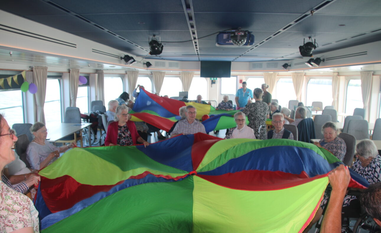 Groep ouderen speelt met een kleurrijke parachute in een ruime zaal met grote ramen, terwijl ze samen plezier hebben.