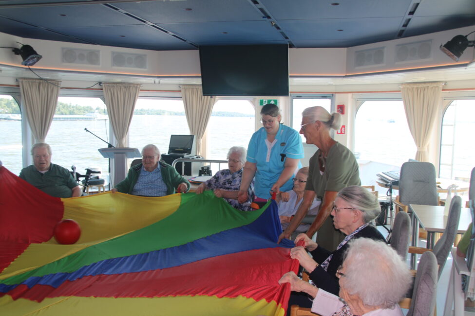 Een groep ouderen speelt met een kleurrijke parachute in een kamer met grote ramen en uitzicht op het water.