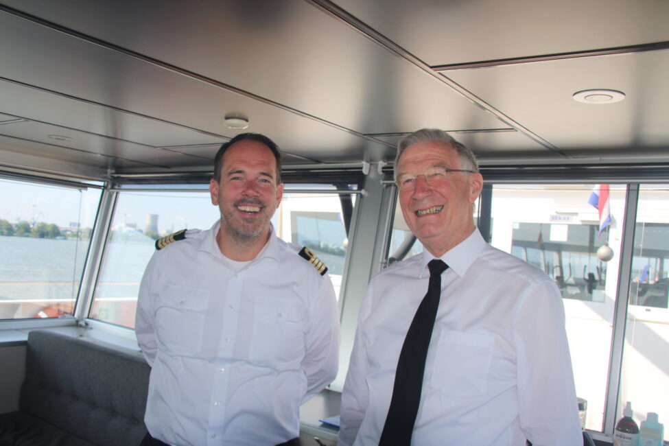Twee mannen in witte overhemden staan lachend op de brug van een schip, met een raam op de achtergrond dat uitzicht biedt op water.