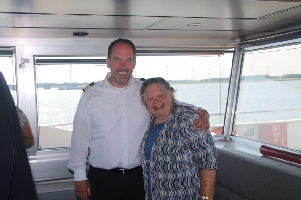 Een man in uniform en een lachende vrouw poseren samen in een boot met grote ramen, met uitzicht op het water op de achtergrond.