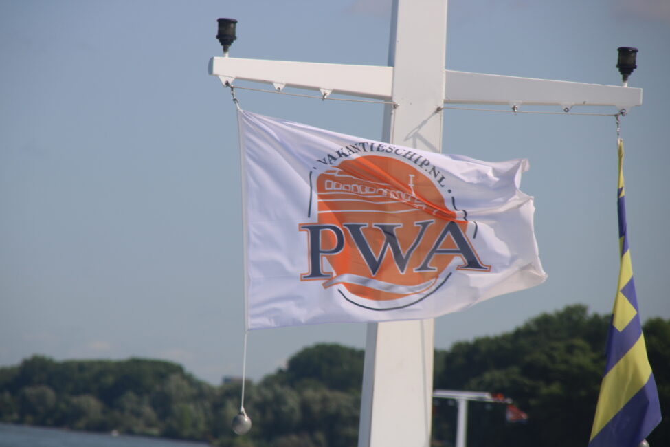Een witte vlag met het logo van PWA wappert aan een mast, met op de achtergrond een blauwe lucht en groene bomen.