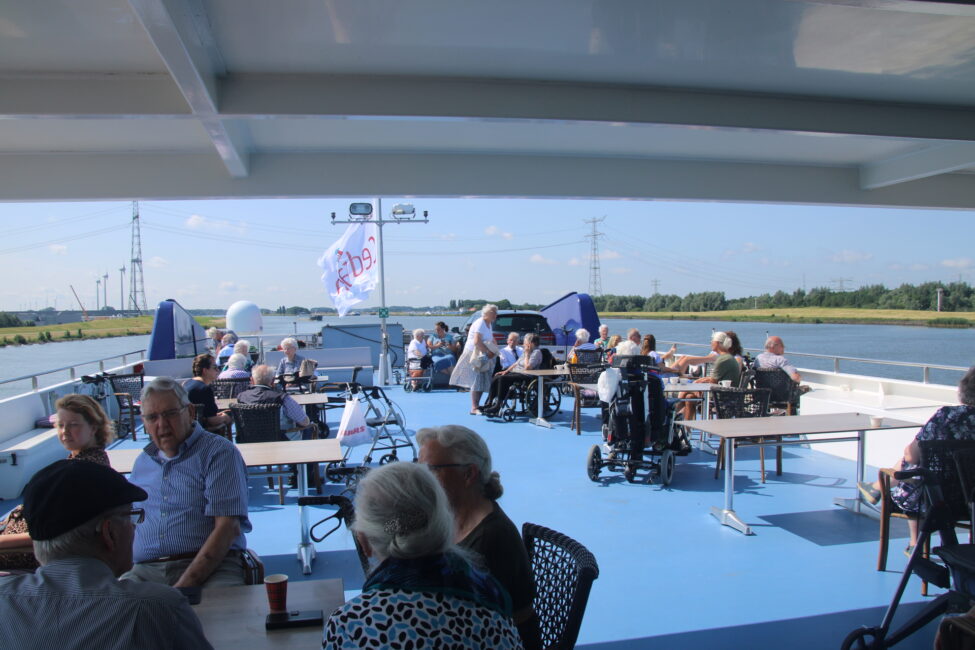 Groep mensen geniet van een zonnige dag op een boot, zittend aan tafels op het dek, met uitzicht op een rivierlandschap.