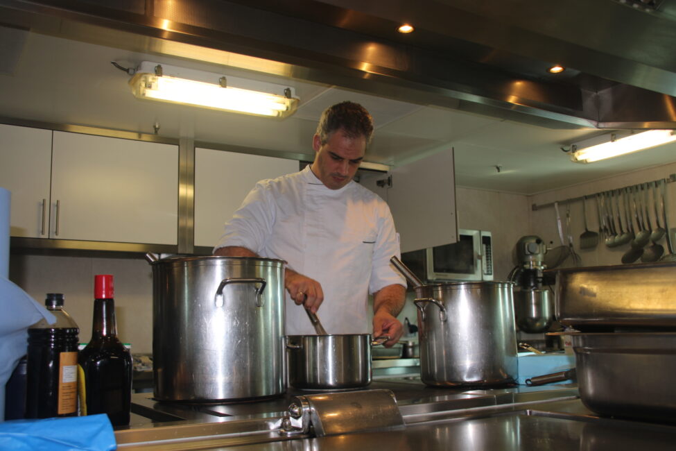 Een kok in een professionele keuken roert in een pan. Omringd door grote potten en flessen, werkt hij geconcentreerd.