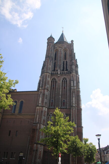 Afbeelding van een hoge, historische kerktoren tegen een blauwe lucht, omringd door groene bomen en een straatlantaarn.