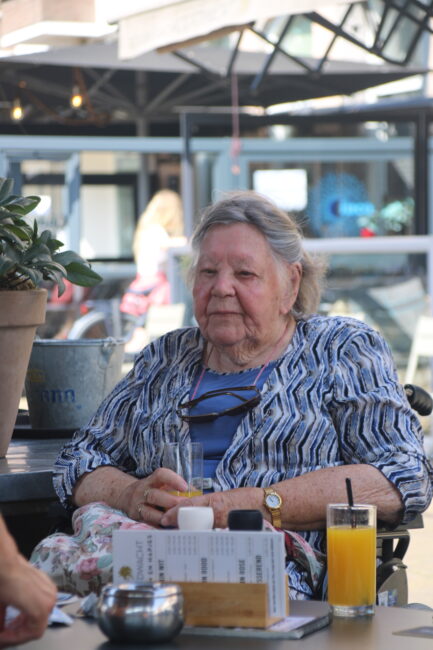Oudere vrouw zit aan een tafel buiten, draagt een blauw-wit gestreepte blouse. Er staat een glas sinaasappelsap op tafel.