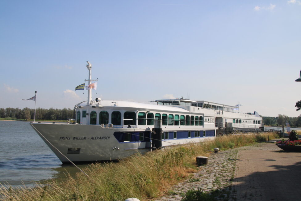 Een riviercruiseschip ligt aangemeerd aan de oever van een rivier, omgeven door gras en een heldere blauwe lucht.