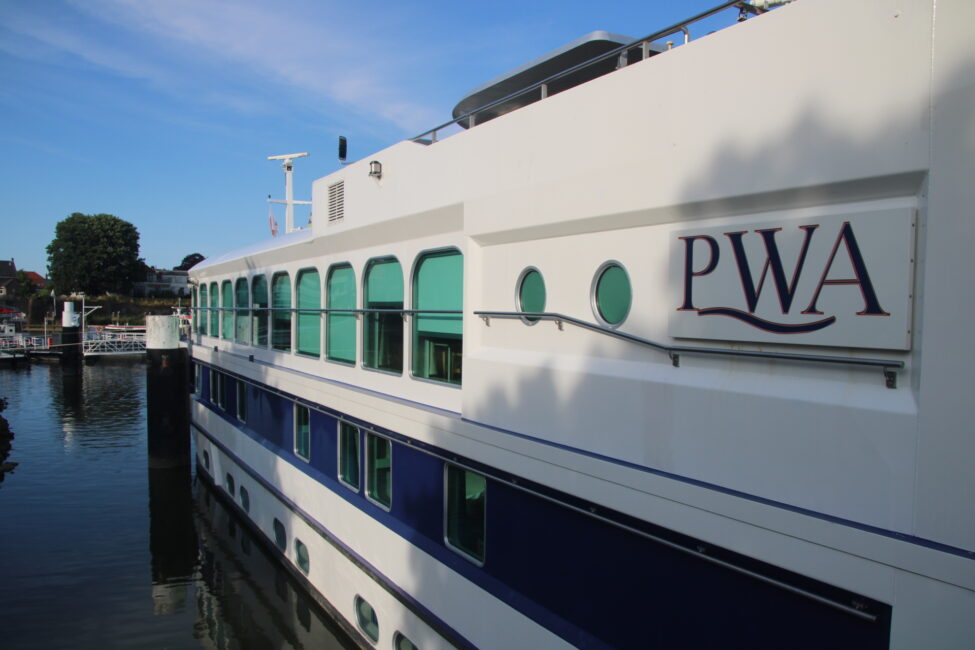 Schip met de letters "PWA" op de zijkant, aangemeerd aan een rustige kade met bomen en een blauwe lucht op de achtergrond.