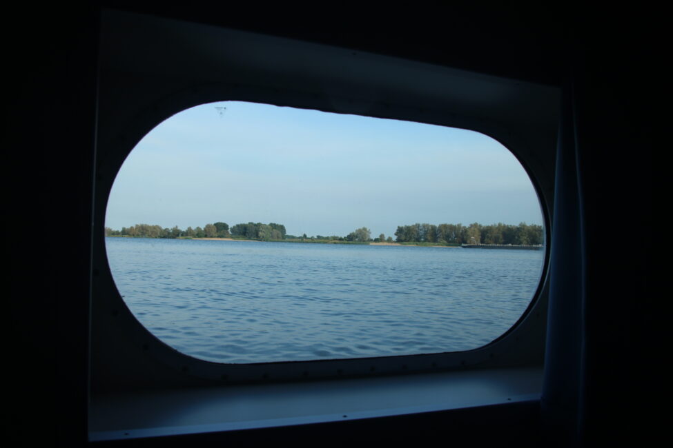 Uitzicht vanuit een scheepsraam op een rustige waterplas met bomen aan de horizon onder een heldere blauwe lucht.
