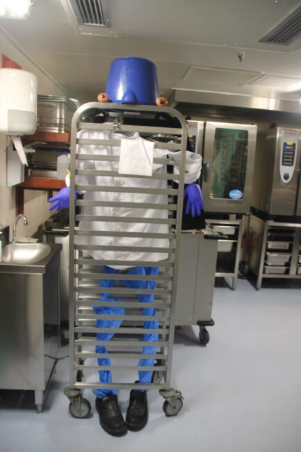Een persoon in een keuken draagt een blauwe emmer op het hoofd en blauwe handschoenen, verstopt achter een metalen rek.