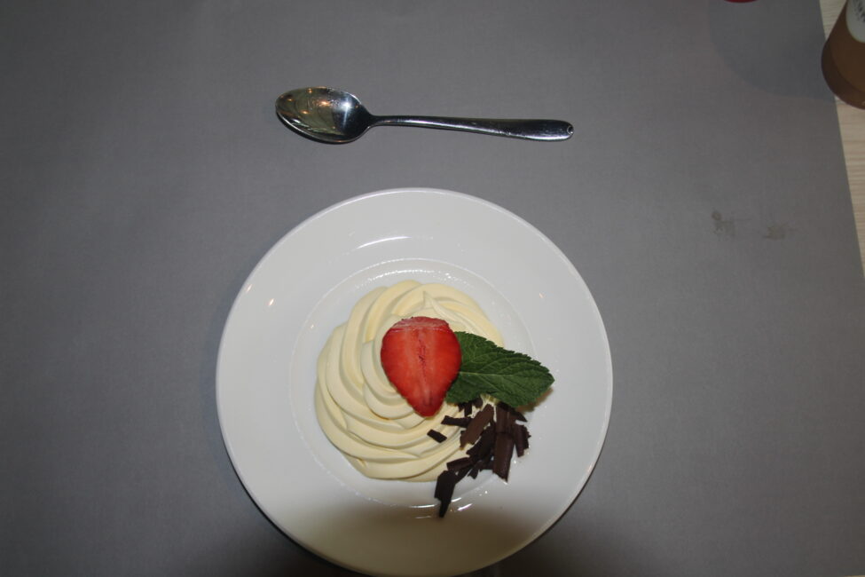 Een wit bord met een dessert van vanillemousse, gegarneerd met een halve aardbei en een muntblaadje, op een grijze ondergrond.