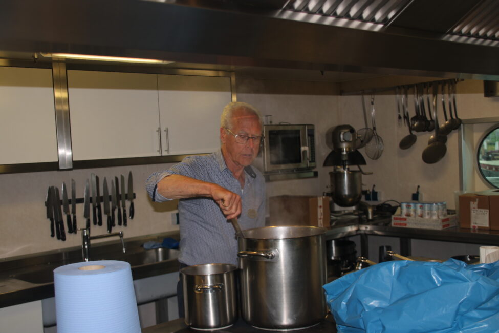 Een oudere man roert in een grote pan in een keuken. Op de achtergrond zijn keukenapparatuur en messen te zien.