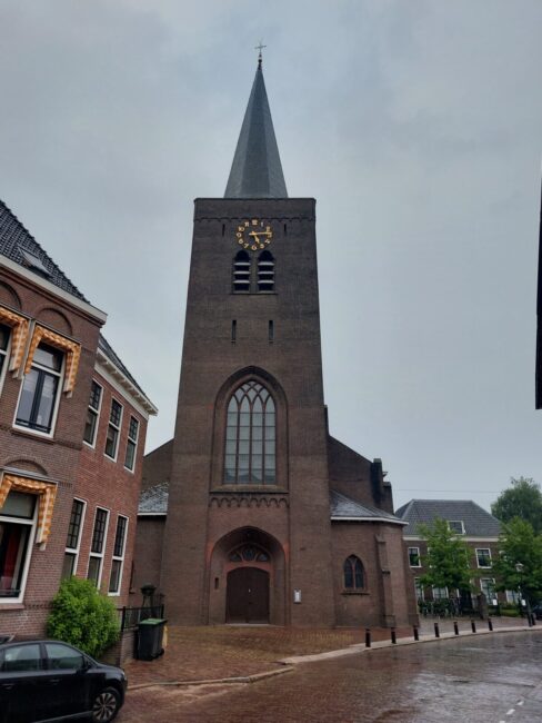 Afbeelding van een historische kerk met een hoge toren en klok, omringd door bakstenen gebouwen op een regenachtige dag.