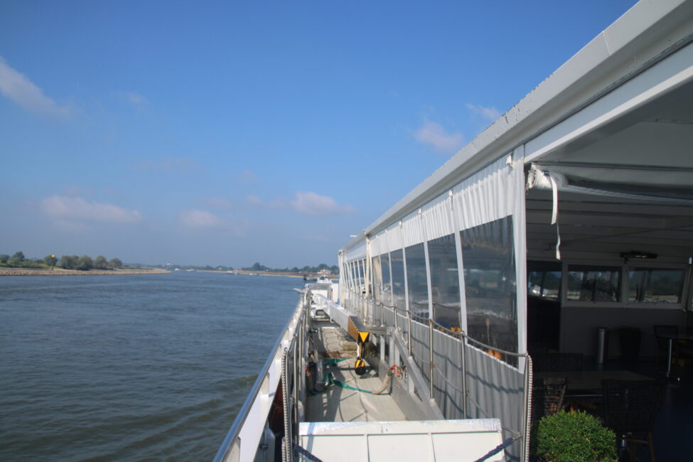 Zijaanzicht van een riviercruiseschip met glazen wanden, varend op een brede rivier onder een heldere blauwe hemel.