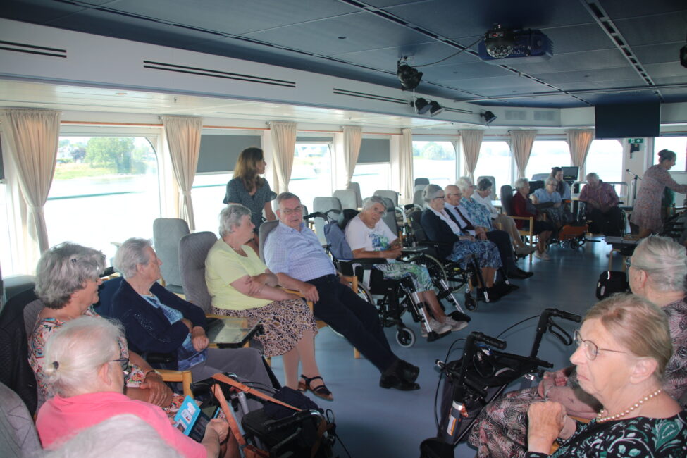 Groep ouderen zit in een salon op een boot, sommigen in rolstoelen. Grote ramen bieden uitzicht op het water en de omgeving.