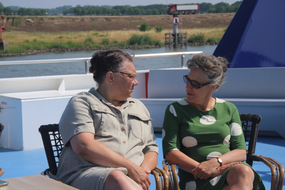 Twee vrouwen zitten buiten op een boot en praten met elkaar. Op de achtergrond is water en een groene oever zichtbaar.