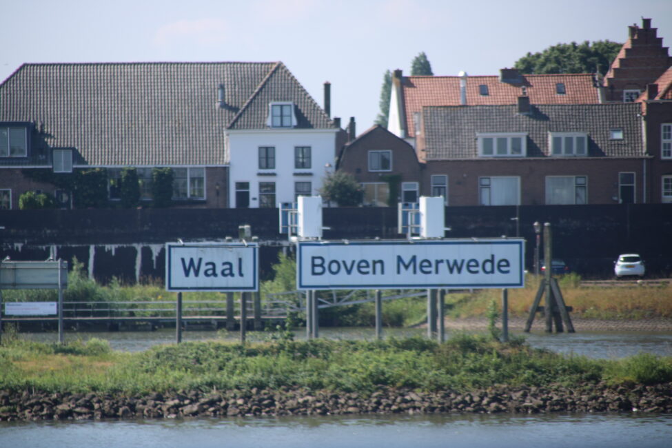 Verkeersborden bij een rivierkruising met de namen "Waal" en "Boven Merwede", op de achtergrond huizen en een groene oever.