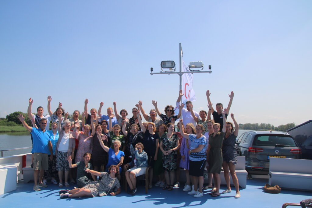 Groep mensen poseert vrolijk op een boot onder een heldere blauwe lucht, met armen in de lucht en een vlag op de achtergrond.