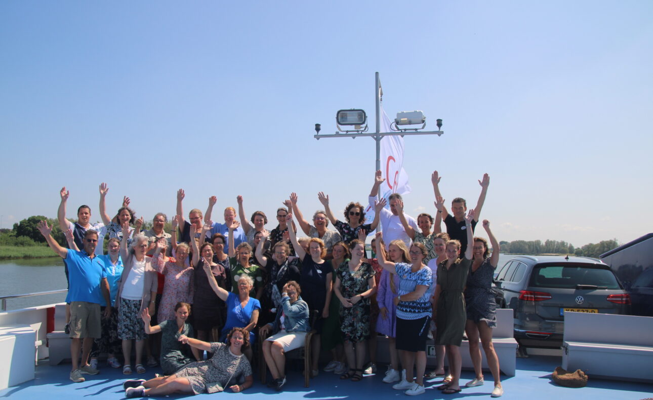 Groep mensen poseert vrolijk op een boot onder een heldere blauwe lucht, met armen in de lucht en een vlag op de achtergrond.