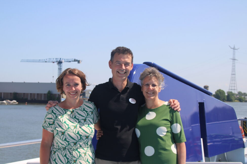 Drie mensen poseren lachend voor een blauwe boot op een zonnige dag. Op de achtergrond zijn een kraan en een waterlichttoren te zien.