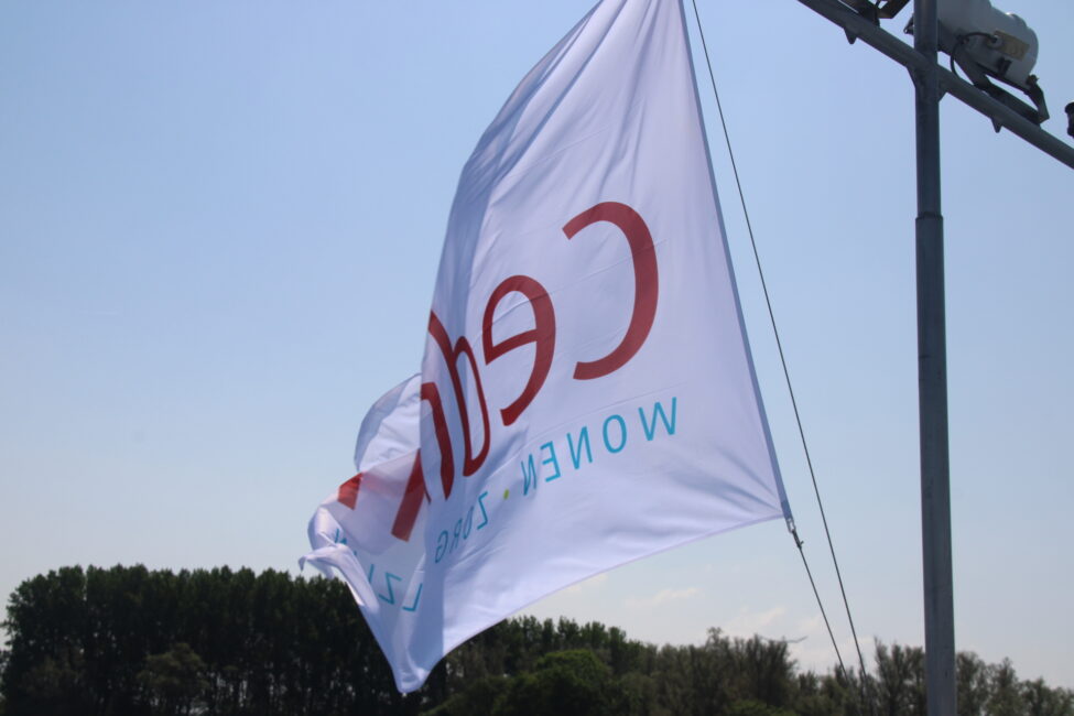 Witte vlag met het rode logo van Cedrah wappert in de wind tegen een blauwe lucht, met bomen op de achtergrond.