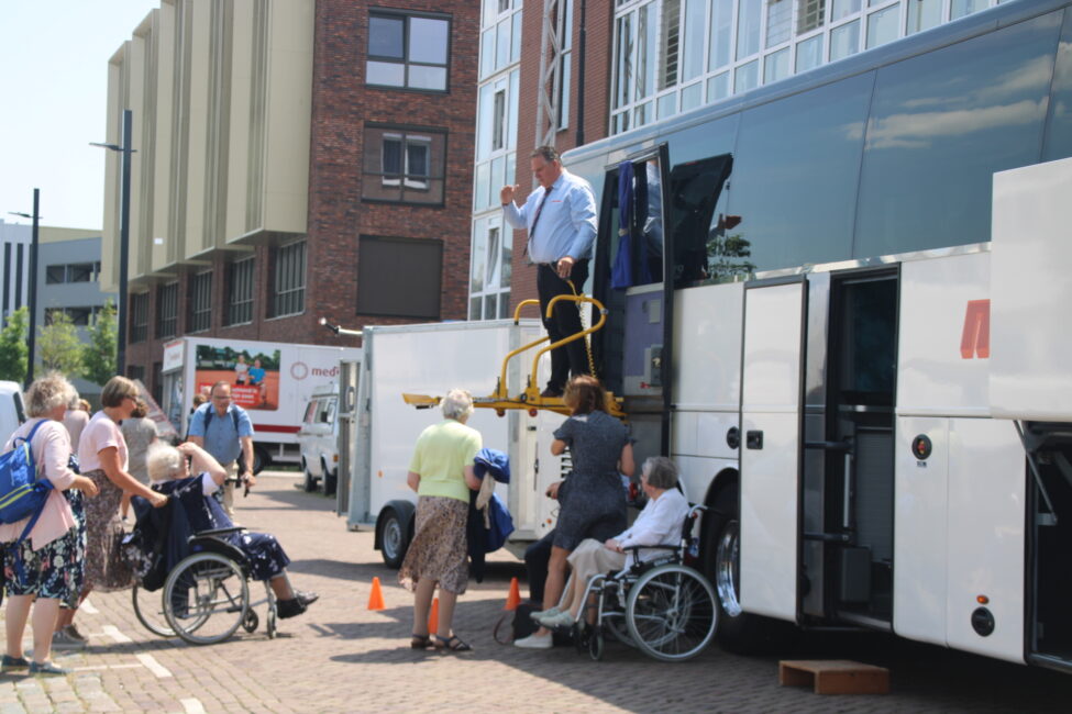 Mensen helpen ouderen in rolstoelen bij het instappen in een bus. Een man staat op een trap en geeft aanwijzingen.