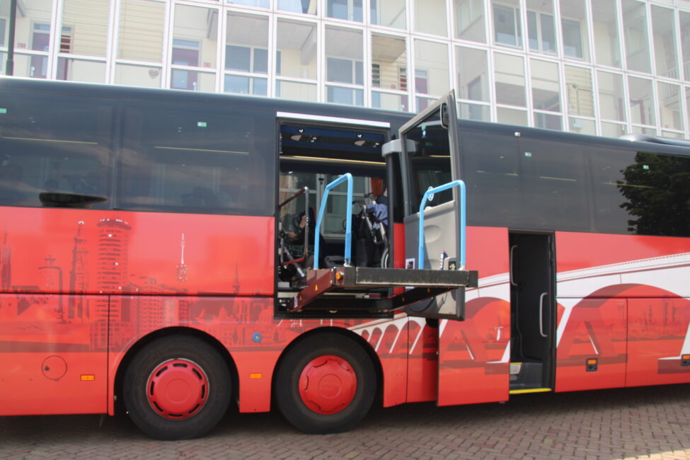 Rode bus met open zijdeur, waar een rolstoellift zichtbaar is. De bus staat geparkeerd voor een gebouw met glazen ramen.