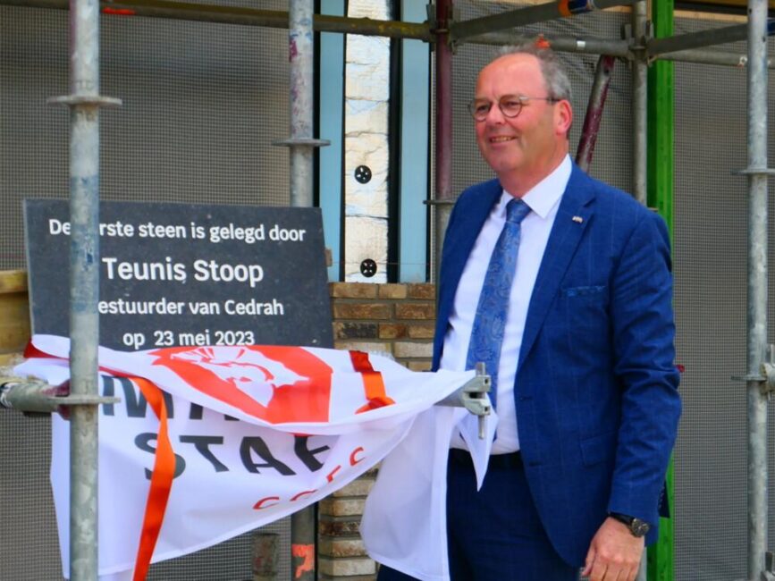 Man in blauw pak onthult een gedenksteen met tekst, omringd door bouwsteigers. Een vlag hangt aan de steiger.