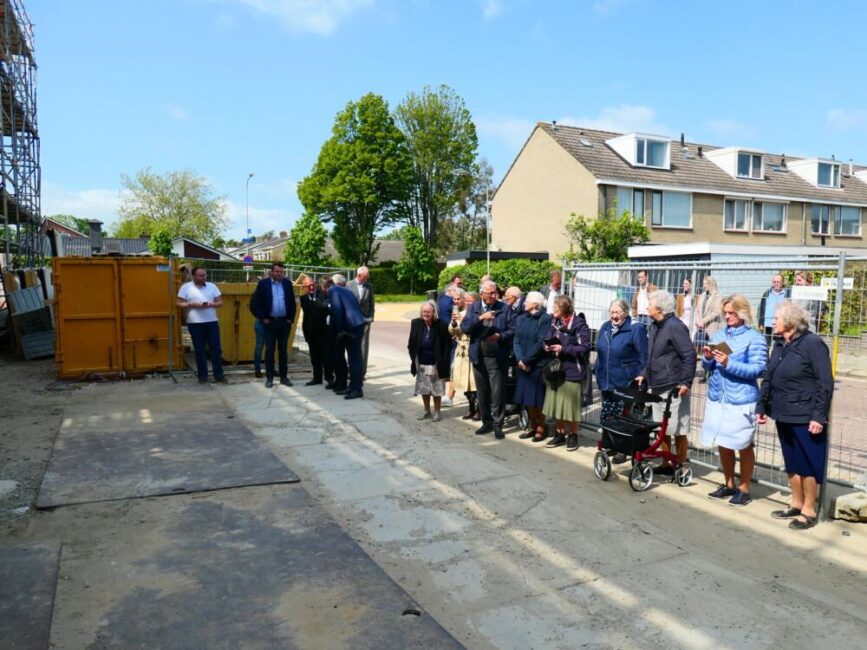 Een groep mensen staat buiten op een zonnige dag, naast een bouwplaats met containers en huizen op de achtergrond.
