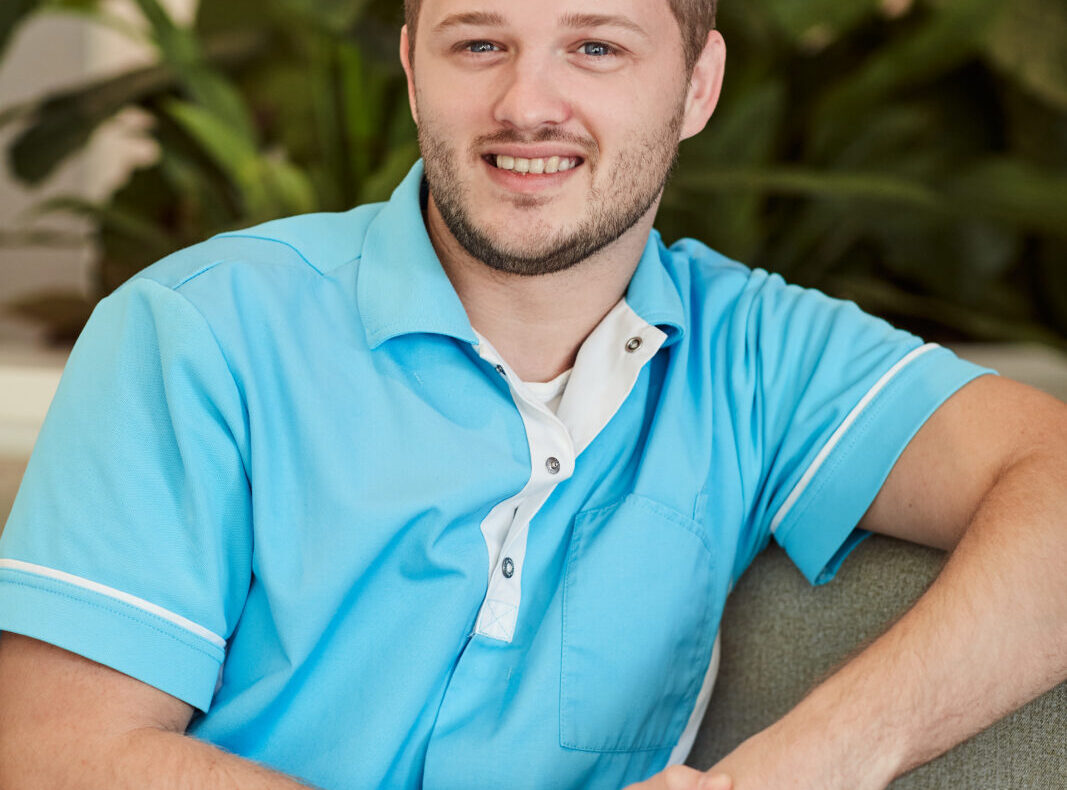Jonge man in een lichtblauw uniform zit glimlachend op een bank, met groene planten op de achtergrond.