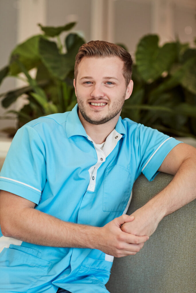 Jonge man in een lichtblauw uniform zit glimlachend op een bank, met groene planten op de achtergrond.