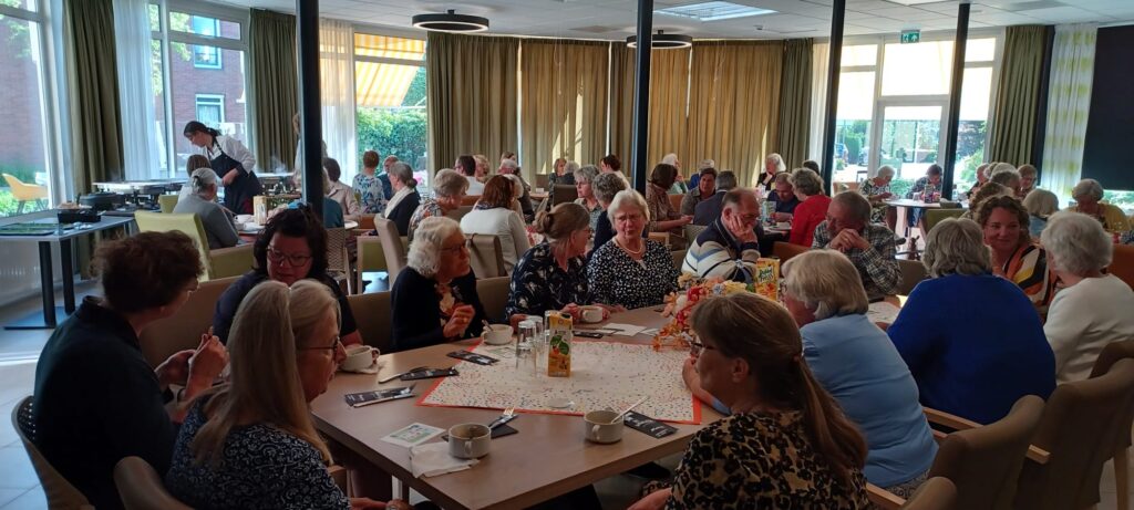 Groep mensen zit aan tafels in een lichte ruimte, genietend van een sociale bijeenkomst met koffie en gebak op tafel.